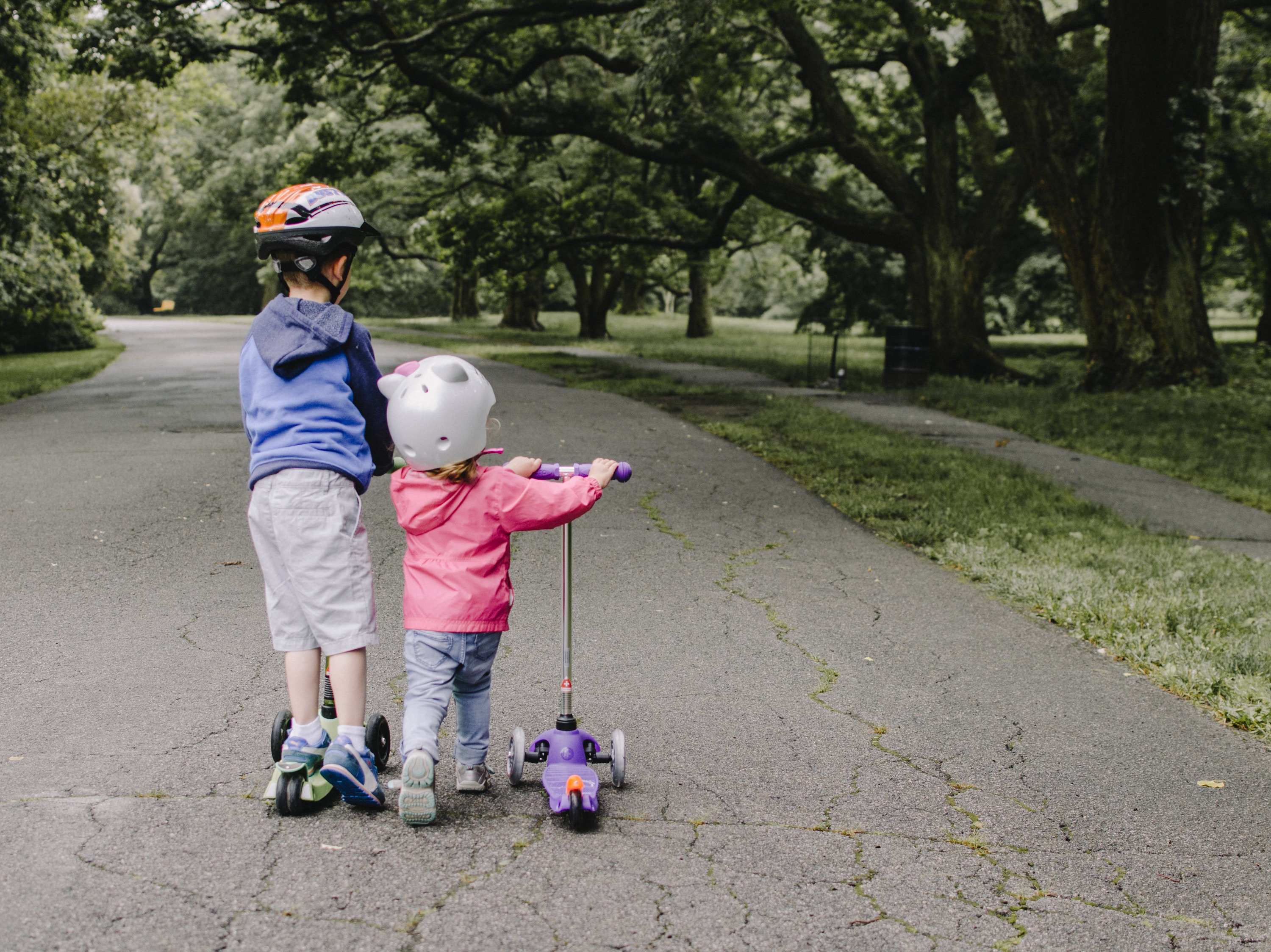 Two young children riding scooters on a park path lined with trees.