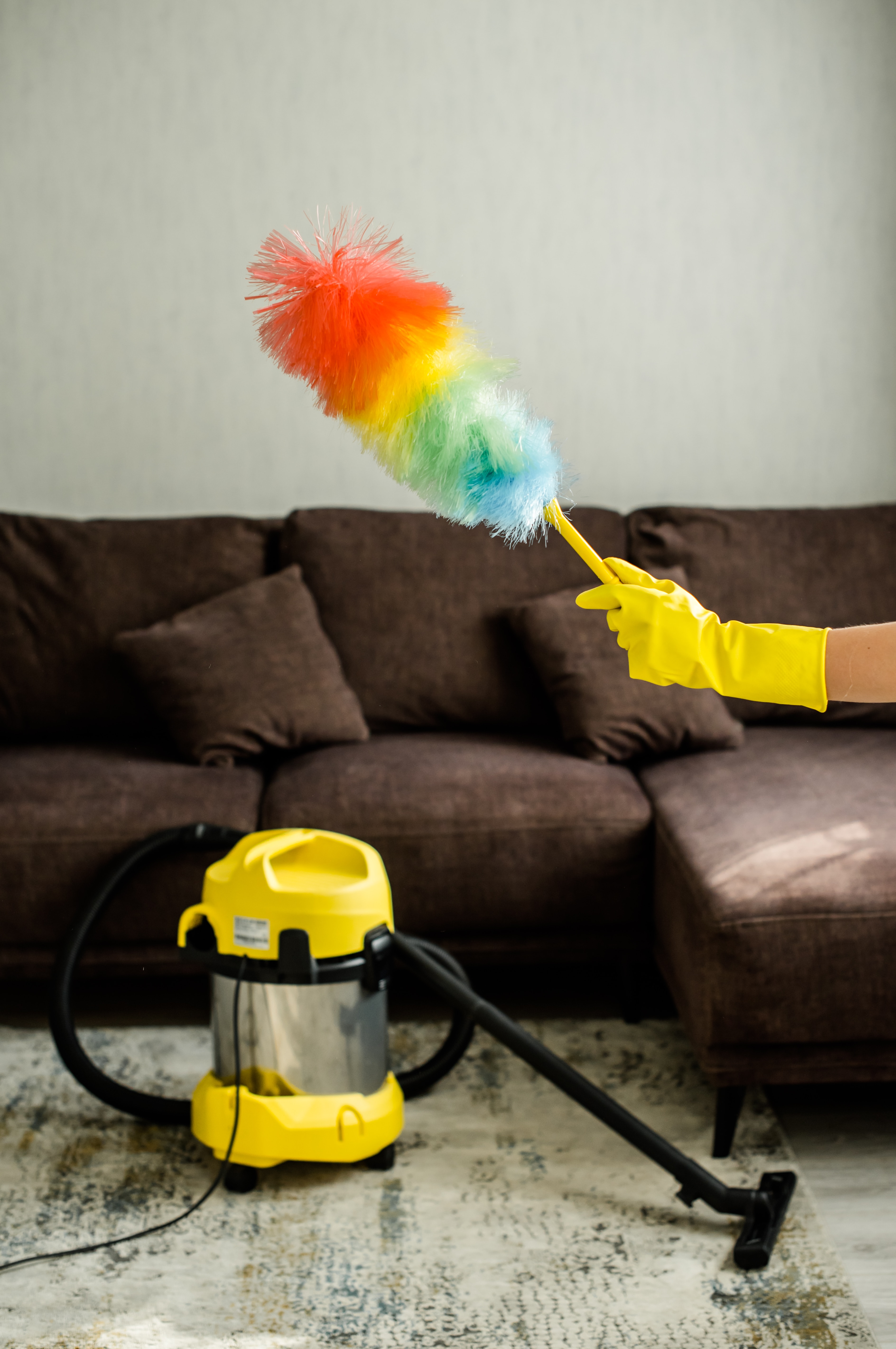 Person using colorful feather duster to clean dark brown couch with wet vacuum below