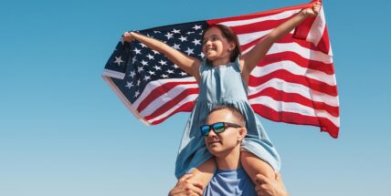 Father holding daughter on shoulders while waving American flag against blue sky