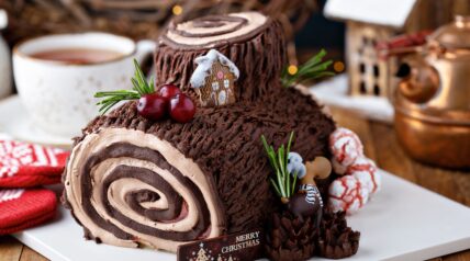Chocolate Yule log cake decorated with gingerbread house, berries, and rosemary sprigs on white